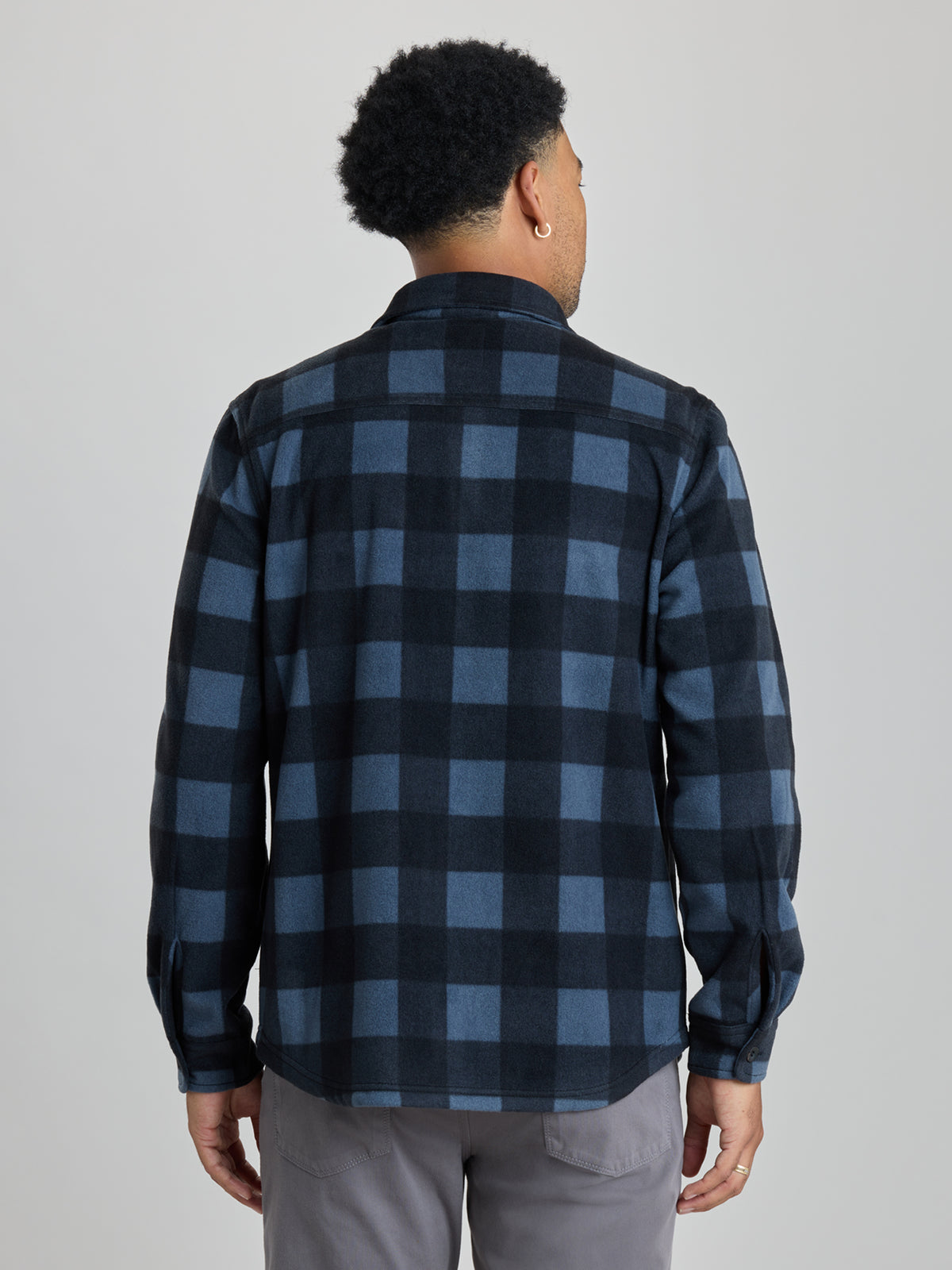 A man viewed from the back wearing a long-sleeve button-up flannel shirt with a dark blue and black buffalo check pattern, paired with gray pants, against a solid light gray background.  | Nightfall