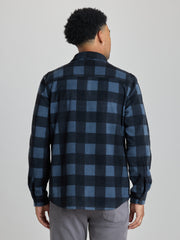 A man viewed from the back wearing a long-sleeve button-up flannel shirt with a dark blue and black buffalo check pattern, paired with gray pants, against a solid light gray background.  | Nightfall