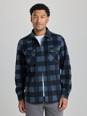 A man wearing a dark blue and black buffalo plaid button-down flannel shirt over a white t-shirt and gray pants, photographed against a solid light gray background. | Nightfall