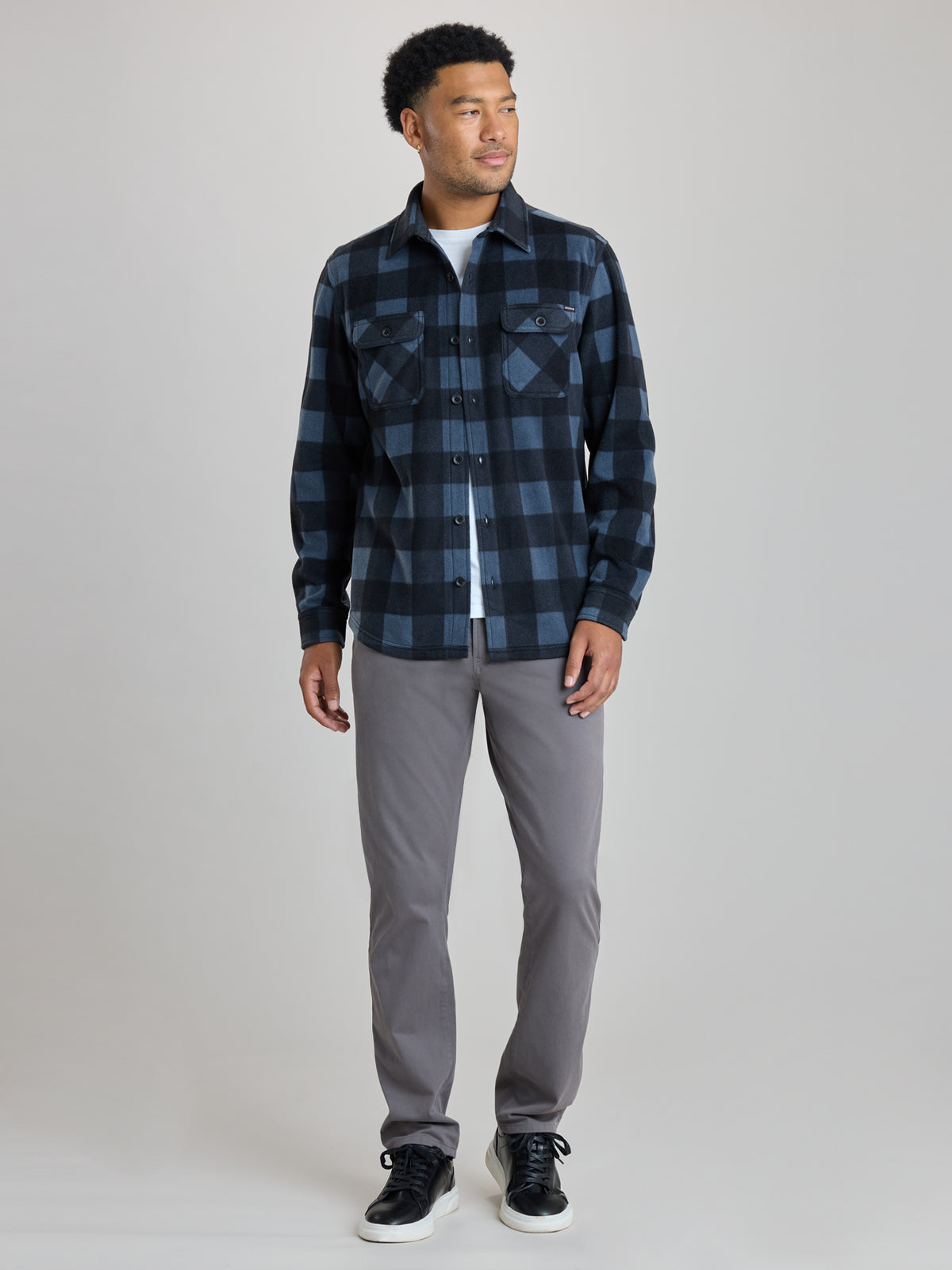 A man modeling a dark blue and black buffalo plaid flannel shirt over a white t-shirt, paired with gray pants and black sneakers, against a light gray studio background.  | Nightfall
