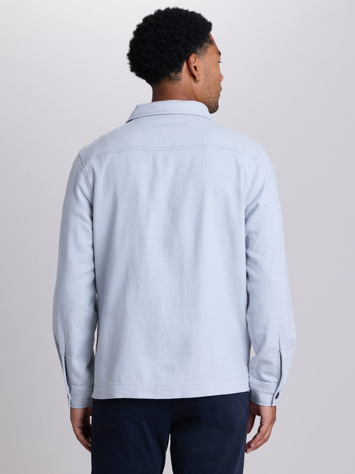A man viewed from the back wearing a light blue long-sleeve button-up shirt and dark navy trousers, set against a plain light gray studio background. | Vapor
