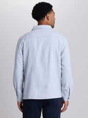 A man viewed from the back wearing a light blue long-sleeve button-up shirt and dark navy trousers, set against a plain light gray studio background. | Vapor