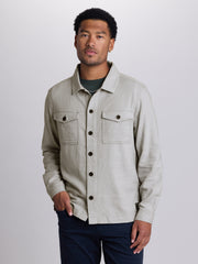 A man wearing a light beige linen button-up shirt jacket over a dark green t-shirt and navy blue pants, standing against a light gray studio background. | Vapor