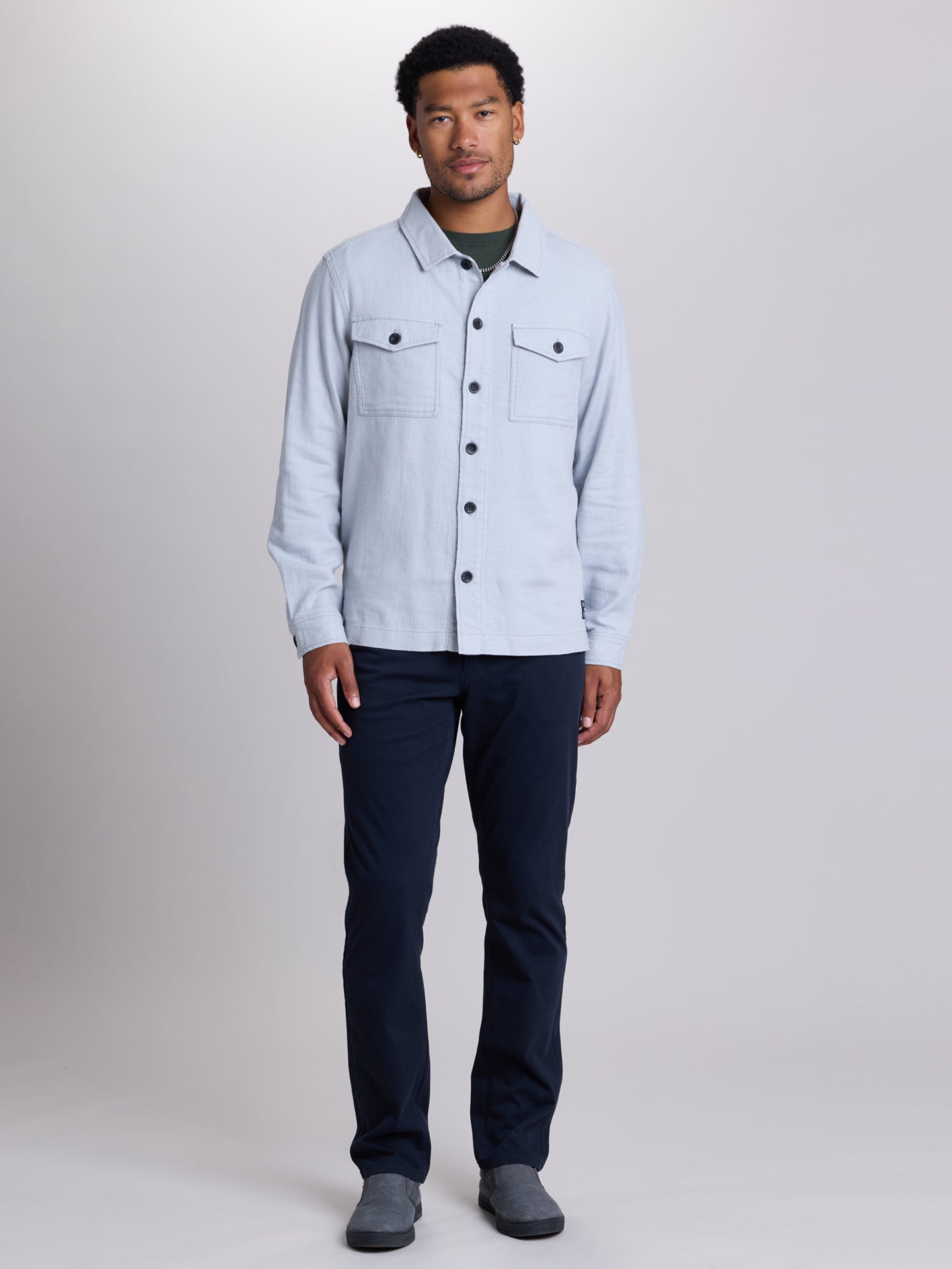 A man modeling a light blue denim shirt jacket over a dark green t-shirt, paired with dark navy pants and gray slip-on shoes, against a neutral gray studio background. | Twilight