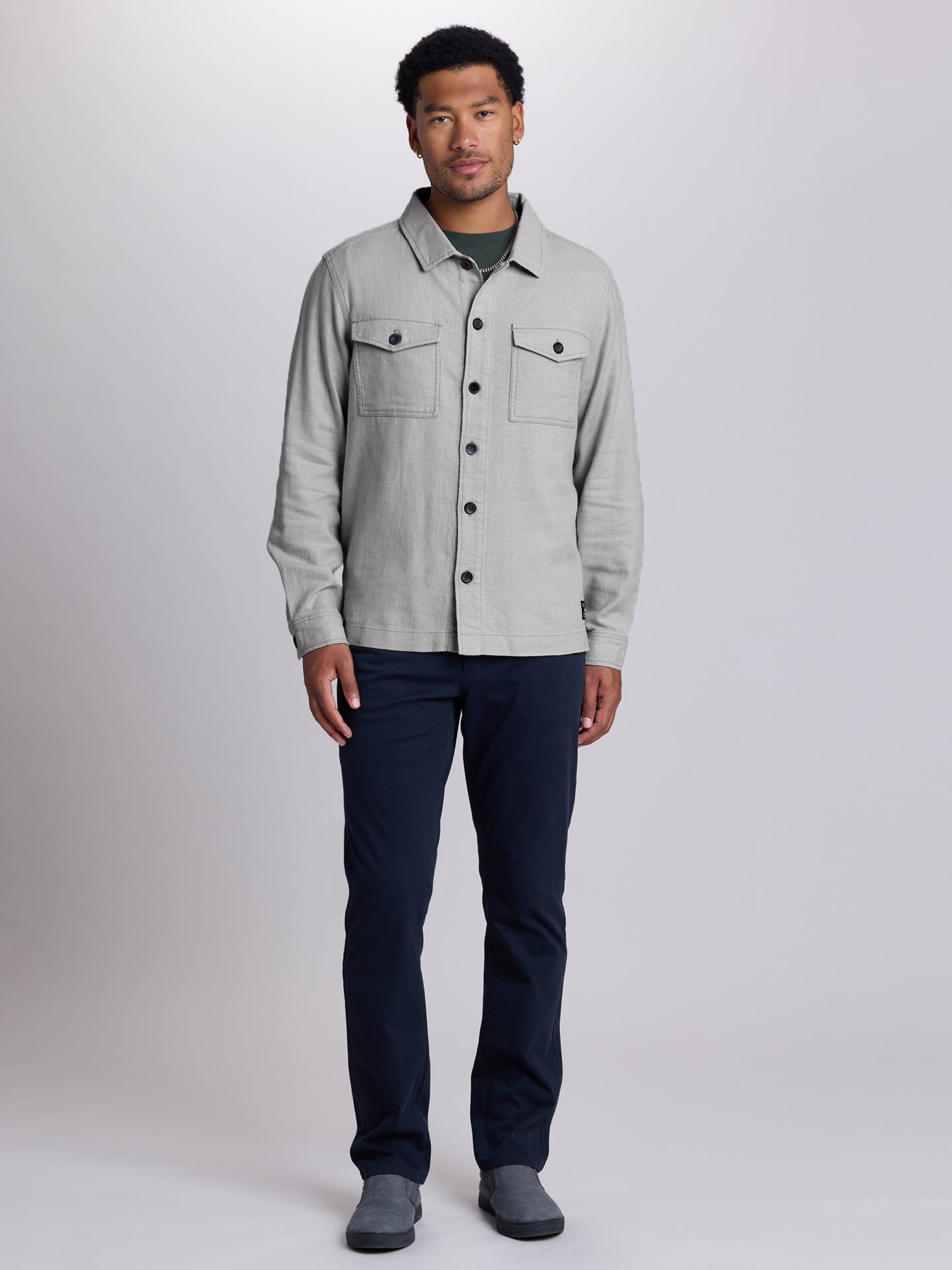 A man modeling a light gray linen button-up shirt jacket over a dark green t-shirt, paired with navy blue pants and gray slip-on shoes, against a light gray studio background. | Vapor