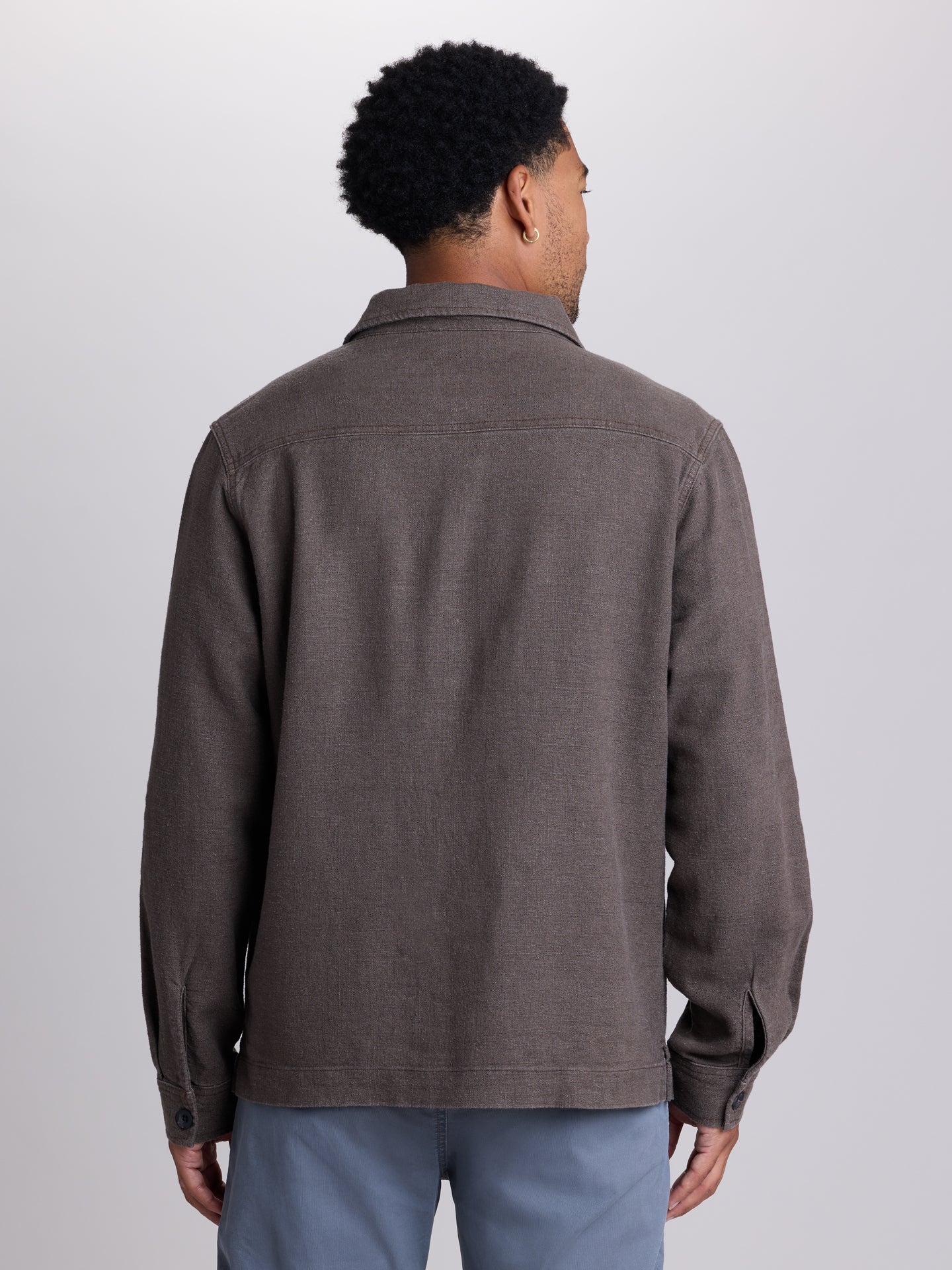 A man viewed from the back wearing a dark brown textured long-sleeve overshirt or lightweight jacket, paired with light blue pants, against a plain light gray studio background. | Carob