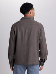 A man viewed from the back wearing a dark brown textured long-sleeve overshirt or lightweight jacket, paired with light blue pants, against a plain light gray studio background. | Carob