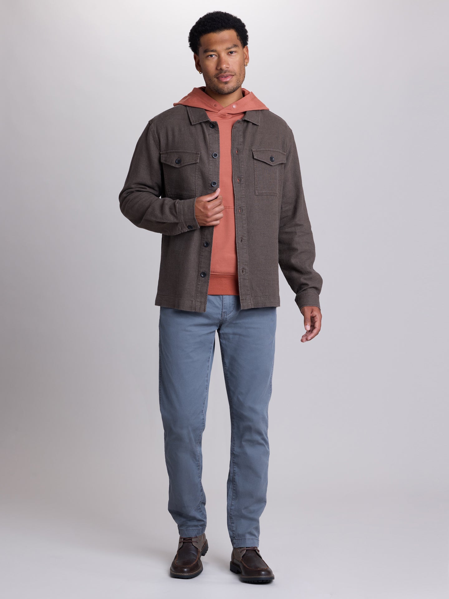 A man modeling a dark brown button-up overshirt jacket over a rust-orange hooded sweatshirt, paired with blue jeans and brown leather shoes, against a light gray studio background. | Carob
