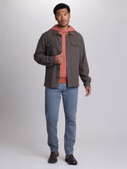A man modeling a dark brown button-up overshirt jacket over a rust-orange hooded sweatshirt, paired with blue jeans and brown leather shoes, against a light gray studio background. | Carob