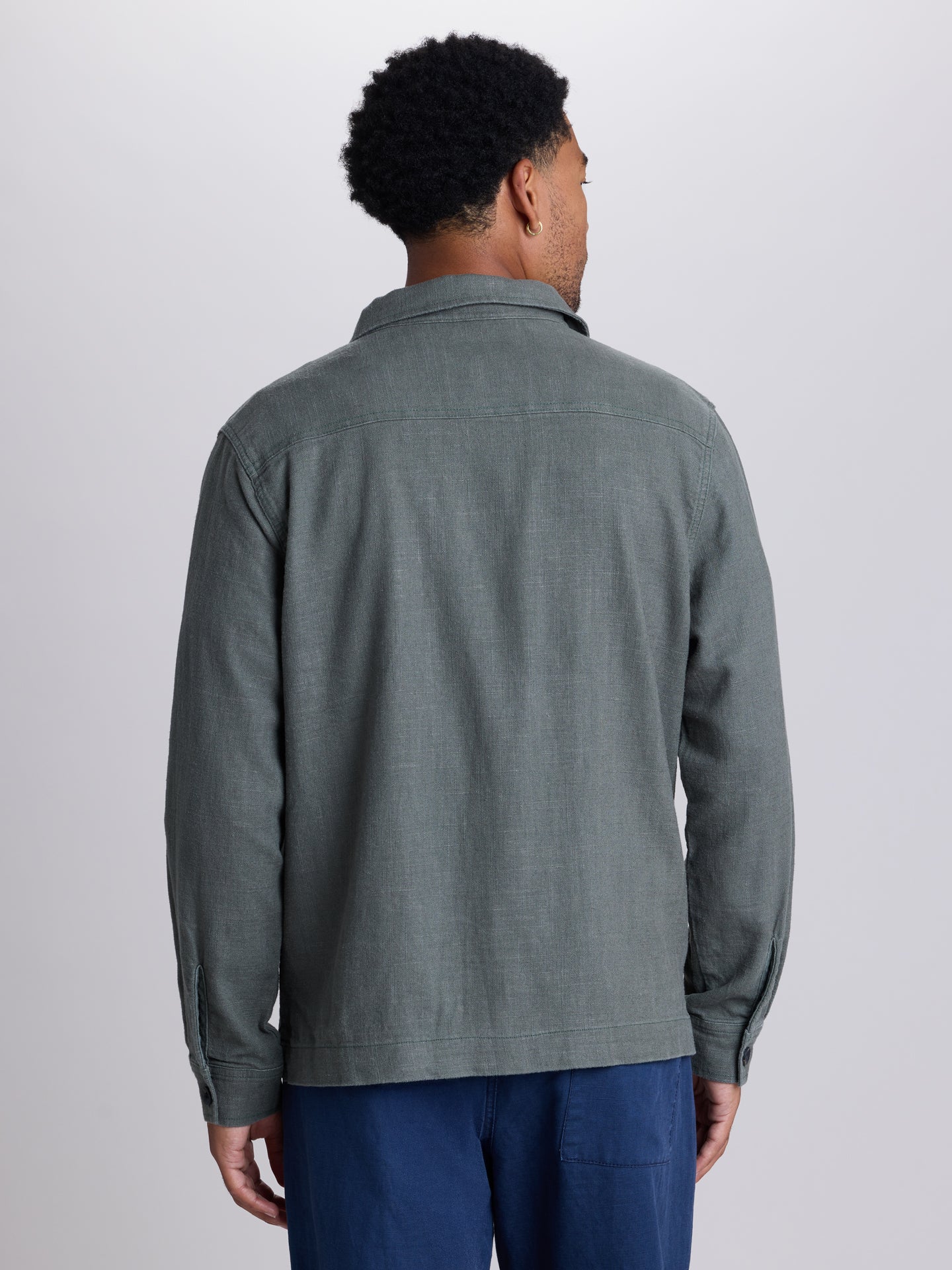 A man viewed from the back wearing a textured green-grey long-sleeve button-up shirt and blue trousers, photographed against a neutral light gray studio background. | Algae