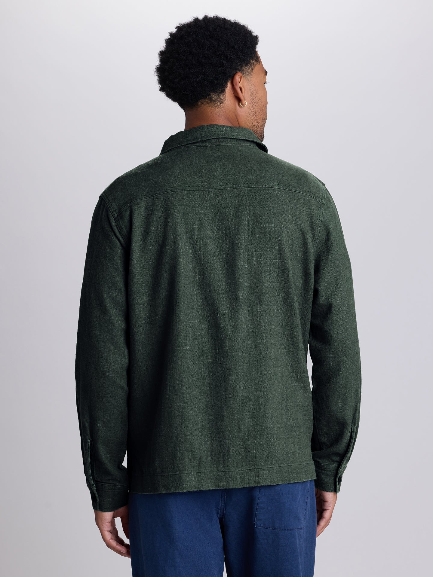 A man viewed from the back wearing a dark green textured long-sleeve shirt and blue pants, standing against a light gray studio background.  | Algae
