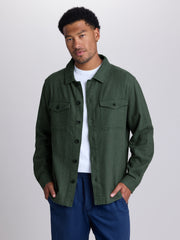 A man wearing an olive green button-up shirt jacket over a white t-shirt and navy blue pants, standing against a light gray studio background. | Algae