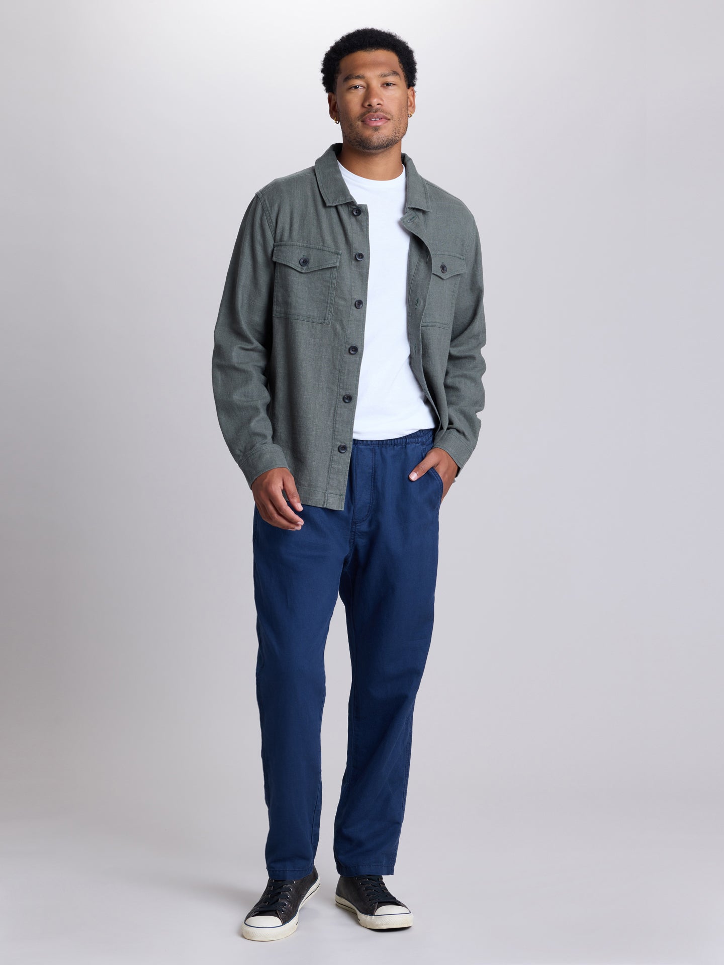 A man modeling a gray button-up overshirt over a white t-shirt, paired with navy blue pants and black sneakers, standing against a light gray studio background. | Deep Blue