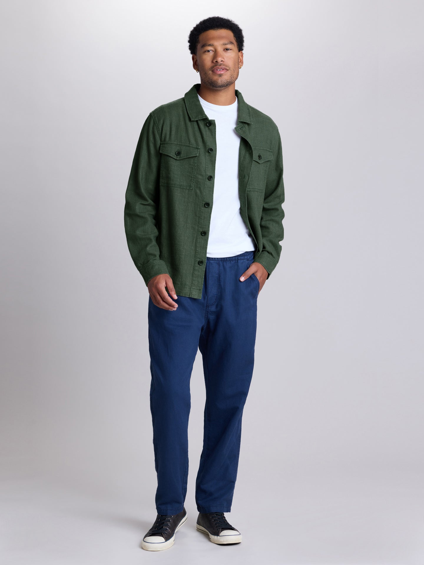 A man modeling a dark green button-up overshirt over a white t-shirt, paired with navy blue pants and black sneakers, standing against a light gray studio background.  | Algae