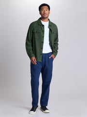 A man modeling a dark green button-up overshirt over a white t-shirt, paired with navy blue pants and black sneakers, standing against a light gray studio background.  | Algae
