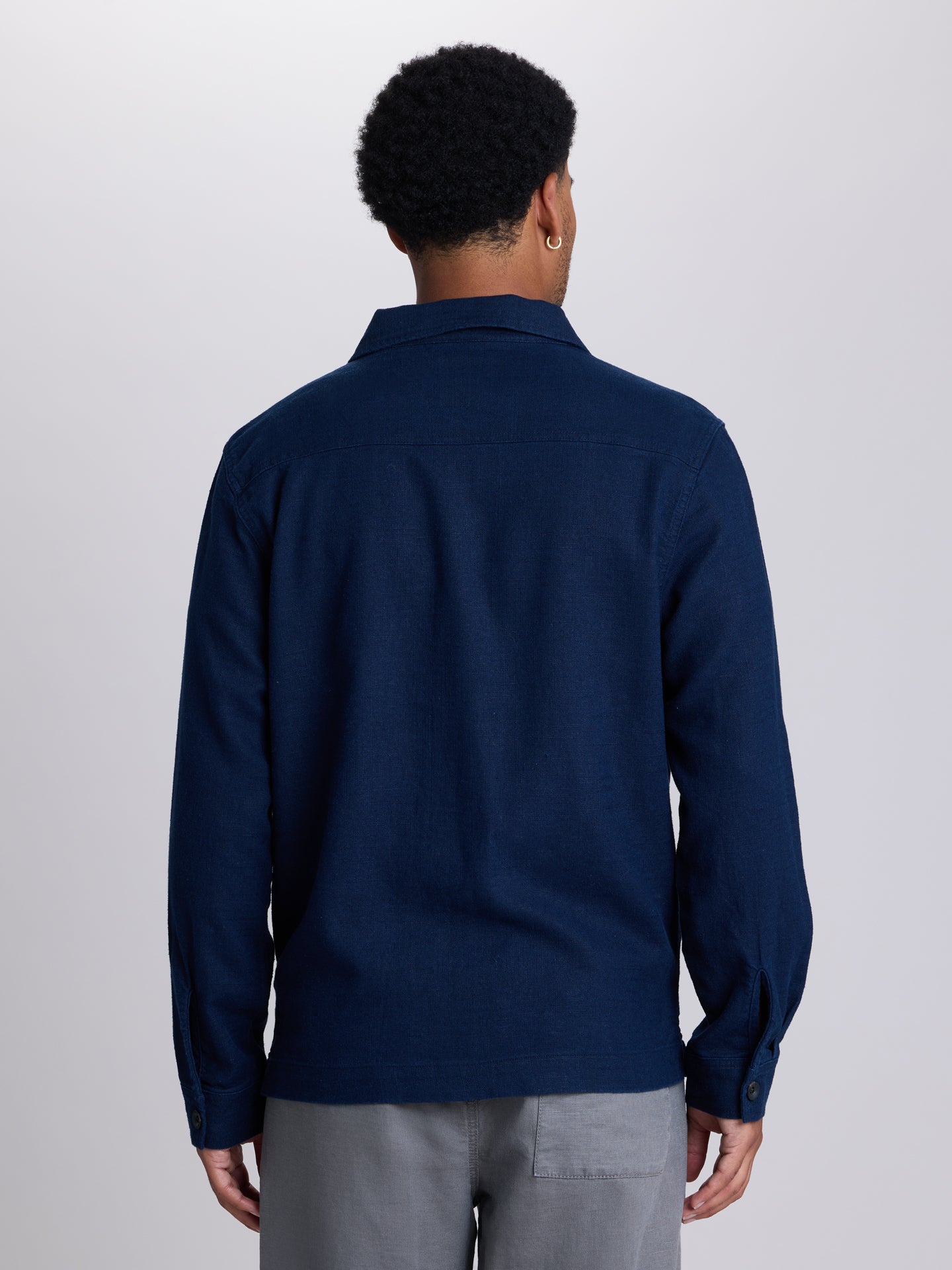 A man viewed from the back wearing a dark navy blue long-sleeve button-up shirt and light gray trousers, set against a plain light gray studio background. | Midnight
