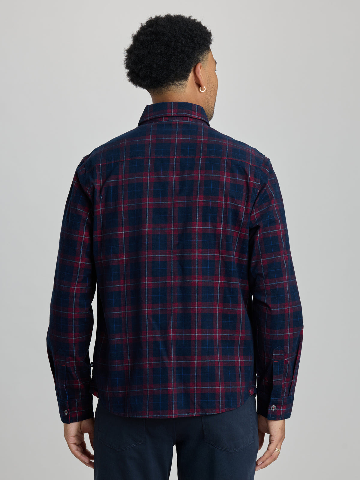 A man viewed from the back wearing a navy blue plaid button-down shirt with red and thin yellow lines, paired with navy pants, against a solid light gray background. | Dark Night