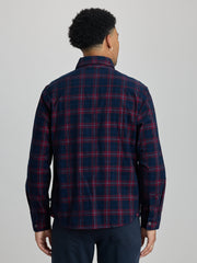 A man viewed from the back wearing a navy blue plaid button-down shirt with red and thin yellow lines, paired with navy pants, against a solid light gray background. | Dark Night
