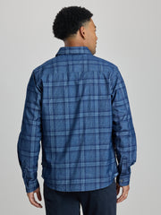 A man viewed from the back wearing a blue plaid button-up shirt and dark pants, standing against a plain light gray studio background. | Blueprint