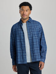 A man wearing a blue and navy plaid button-down shirt over a light gray t-shirt and dark pants, standing against a solid light gray background. | Blueprint