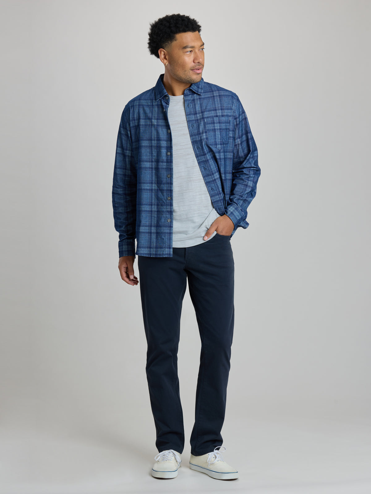 A man modeling a blue plaid button-down shirt over a light gray t-shirt, paired with dark navy pants and white sneakers, against a solid light gray studio background. | Blueprint