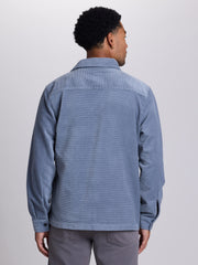 A man viewed from the back wearing a light blue corduroy button-up shirt and gray trousers, photographed against a plain light gray studio background. | Blue Shadow