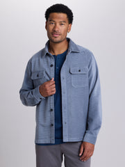 A man wearing a light blue corduroy button-up overshirt over a navy t-shirt and gray pants, standing against a neutral light gray studio background. | Blue Shadow