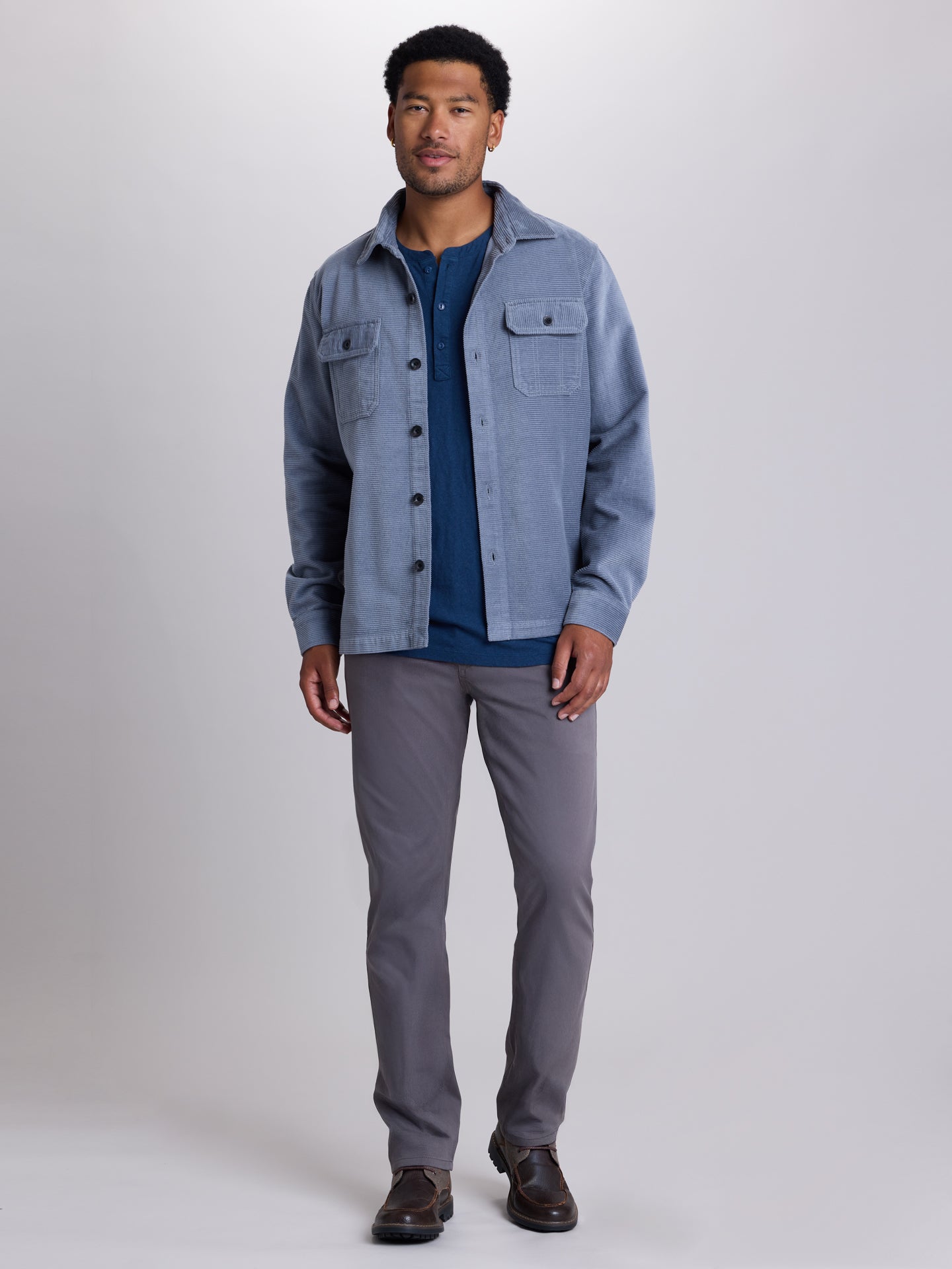 A man modeling a light blue corduroy button-up shirt jacket over a navy blue t-shirt, paired with gray trousers and brown leather shoes, against a light gray studio background. | Blue Shadow