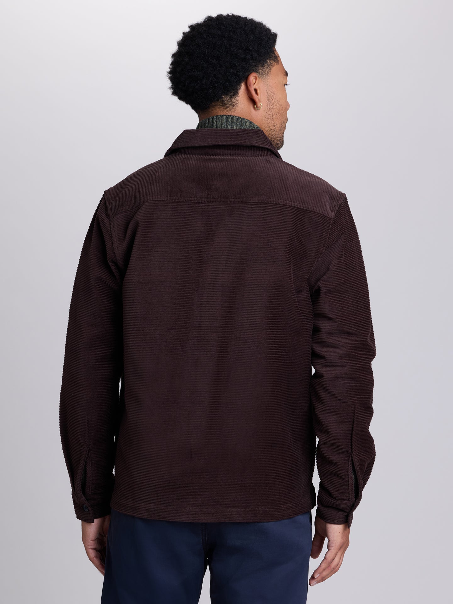 A man viewed from the back wearing a dark brown corduroy jacket over a green patterned shirt and navy pants, set against a plain light gray studio background.  | Lost