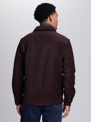 A man viewed from the back wearing a dark brown corduroy jacket over a green patterned shirt and navy pants, set against a plain light gray studio background.  | Lost