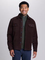 A man wearing a dark brown corduroy button-up shirt jacket over a dark green quarter-zip knit sweater and navy blue jeans, photographed against a neutral light gray studio background. | Lost