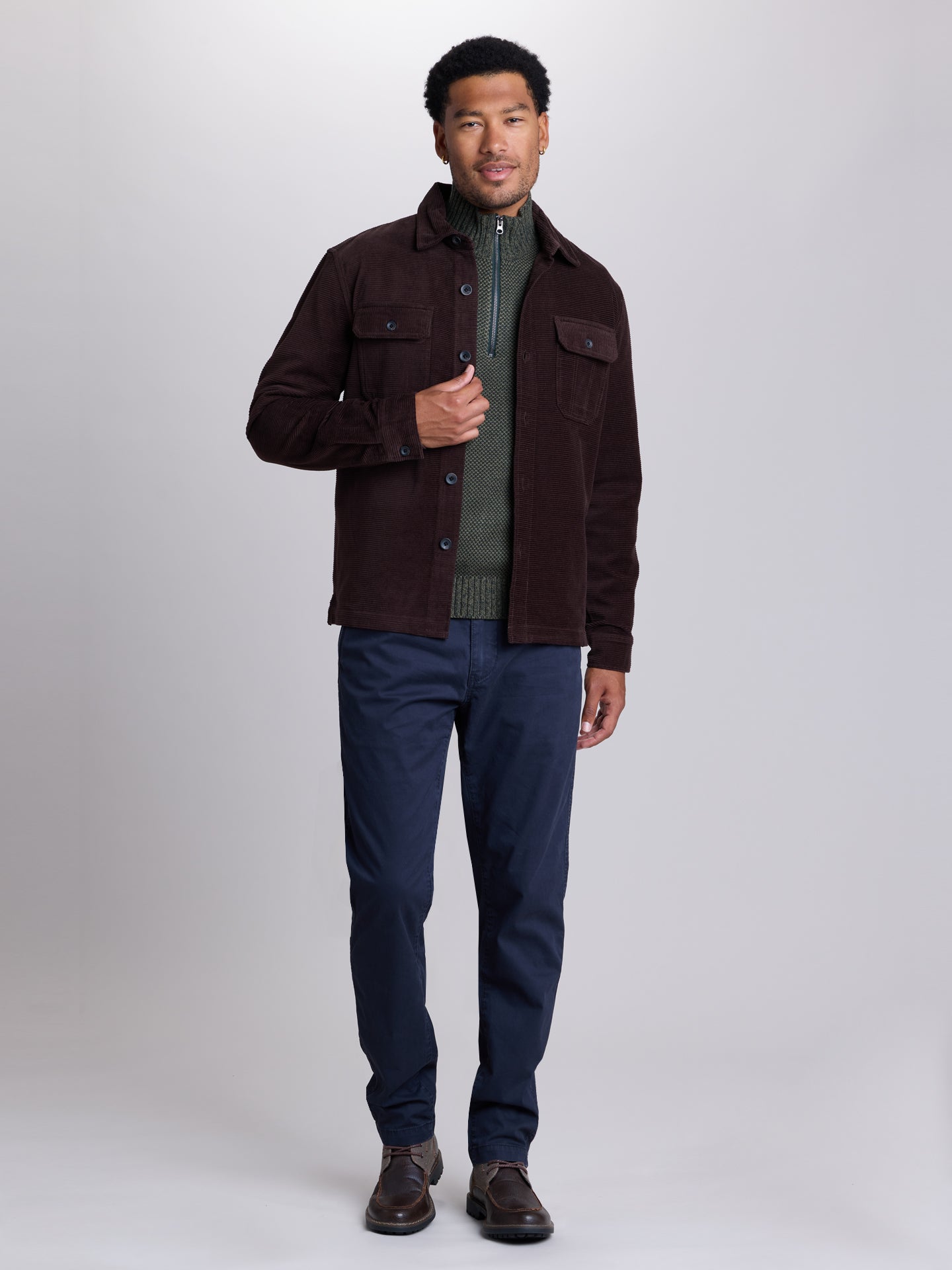 A man modeling a dark brown corduroy jacket over a green ribbed shirt, paired with navy blue pants and brown leather shoes, against a light gray studio background.  | Lost