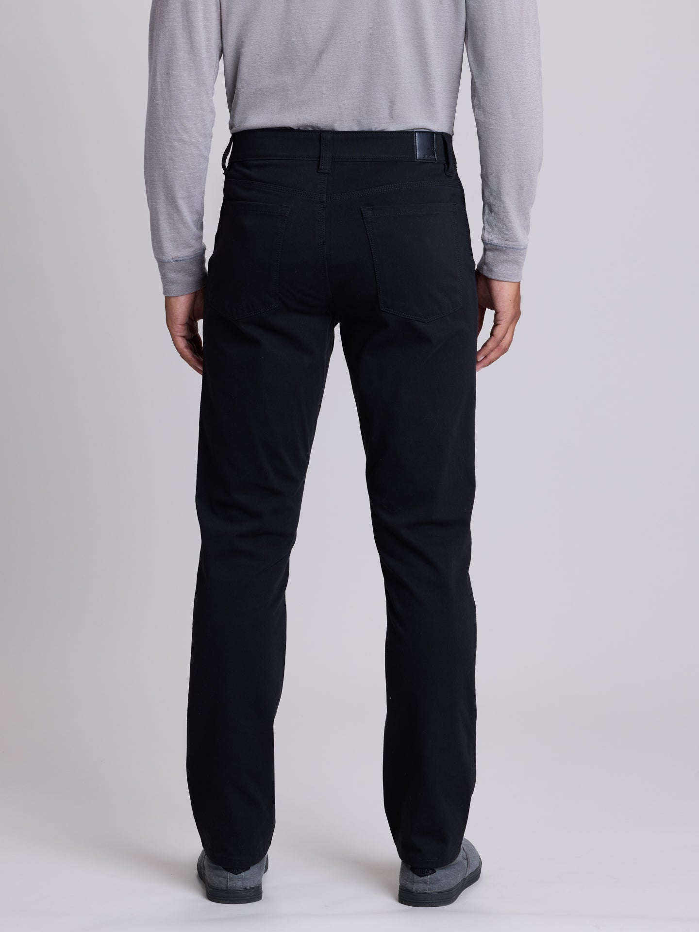 Rear view of a person wearing black trousers, a light gray long-sleeve shirt, and gray shoes against a plain light gray studio background. | Black