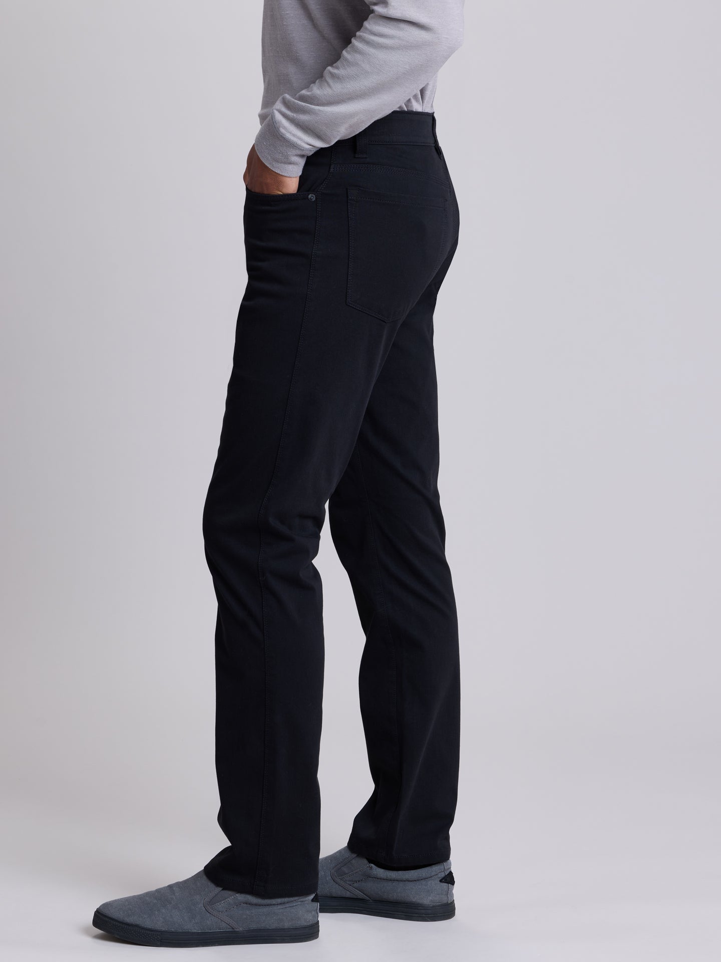 A side-profile view of a person wearing black slim-fit trousers, a light gray long-sleeve top, and dark gray slip-on shoes against a light gray studio background. | Black