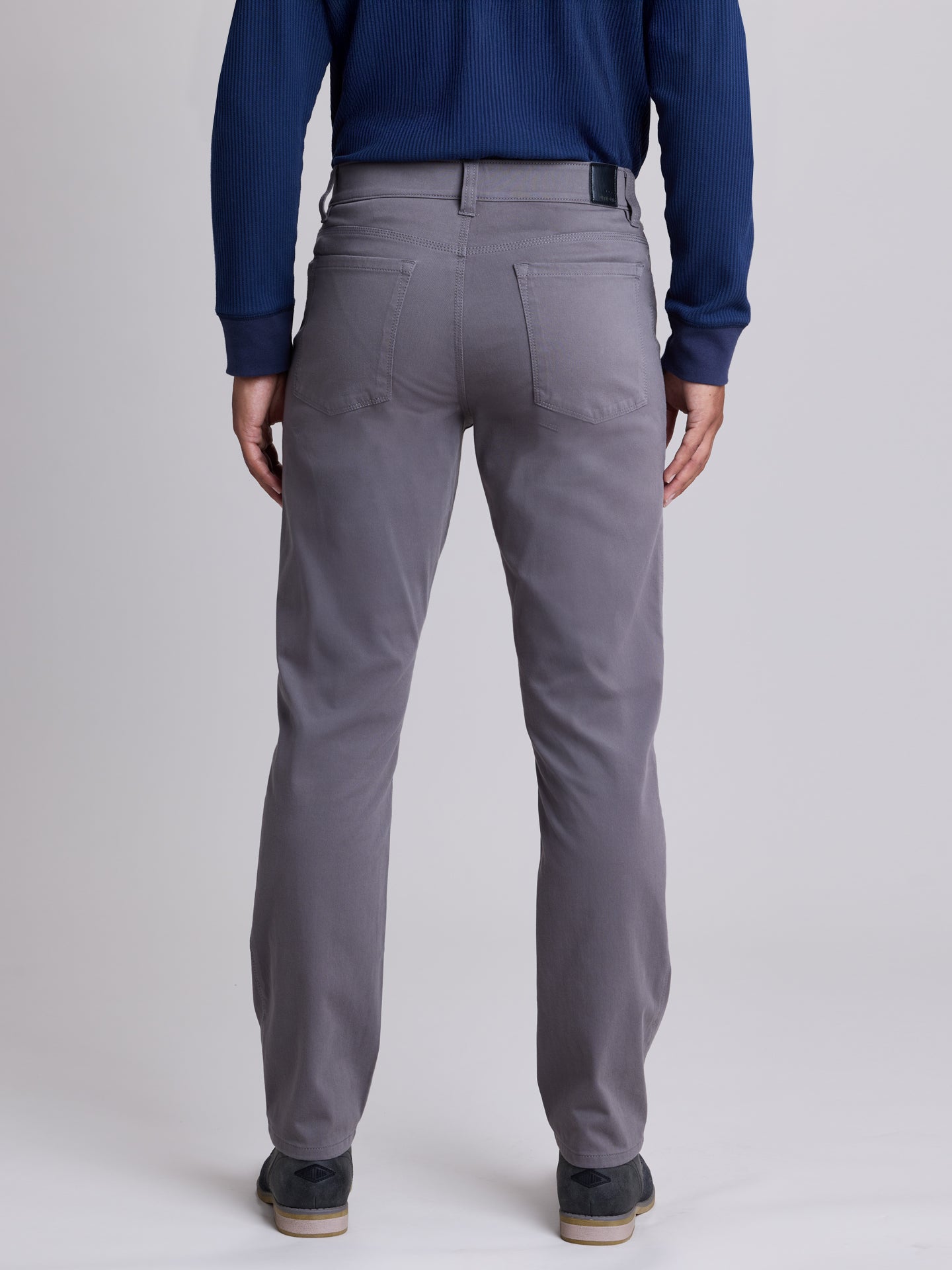 Rear view of a person wearing gray slim-fit trousers, a navy blue ribbed sweater, and dark shoes against a light gray studio background. | Grey Goose
