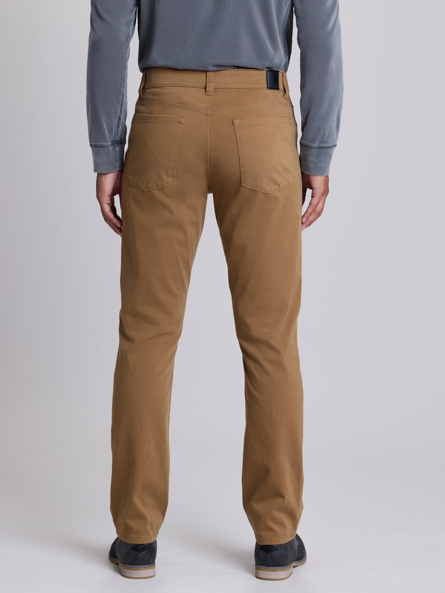 A person shown from the back wearing straight-leg tan chinos with a blue-gray long-sleeve shirt and dark shoes, photographed against a light gray studio background. | Chestnut