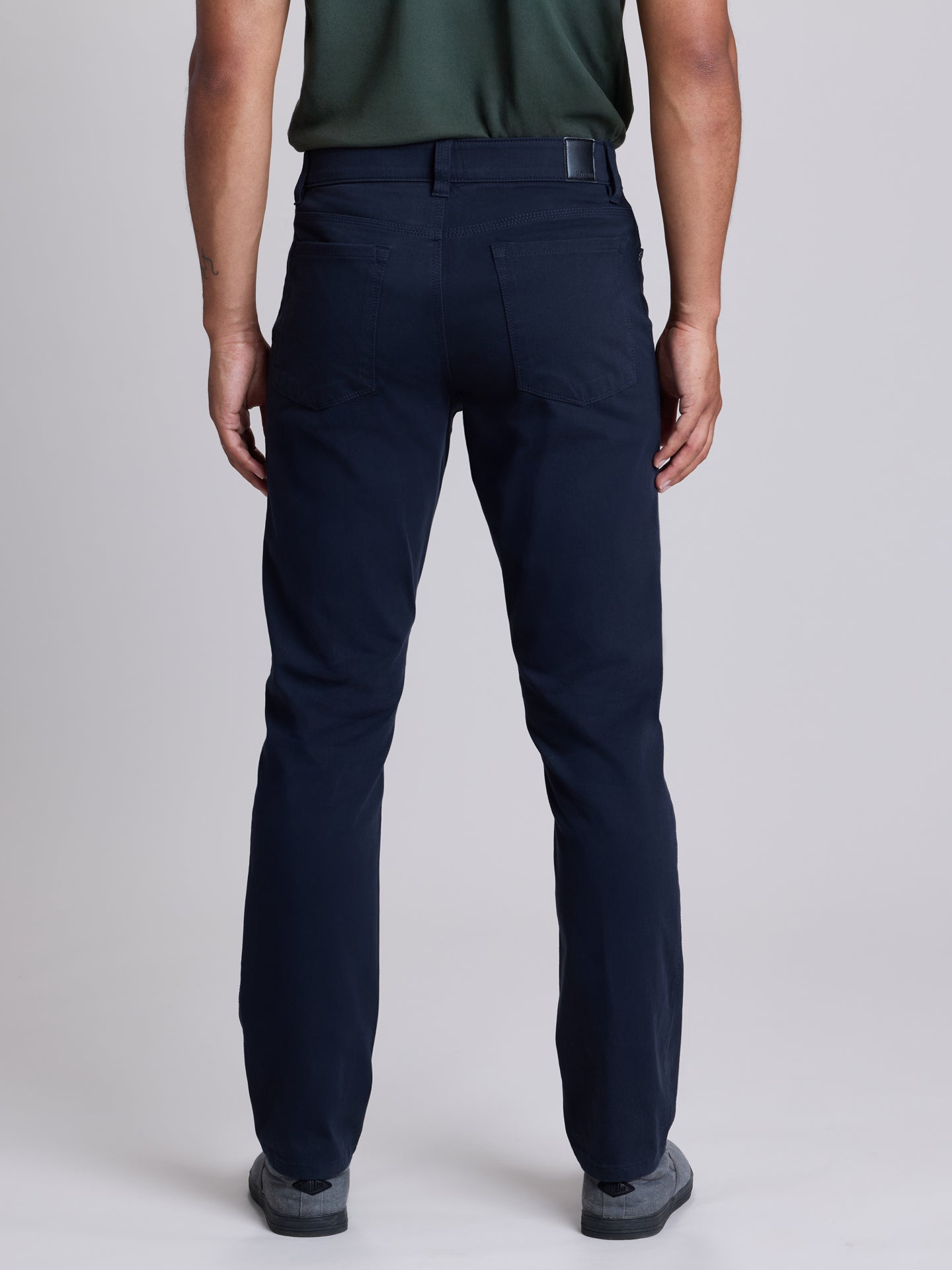 Rear view of a person wearing dark navy straight-leg trousers, a dark green shirt, and gray shoes against a light gray studio background. | Twilight