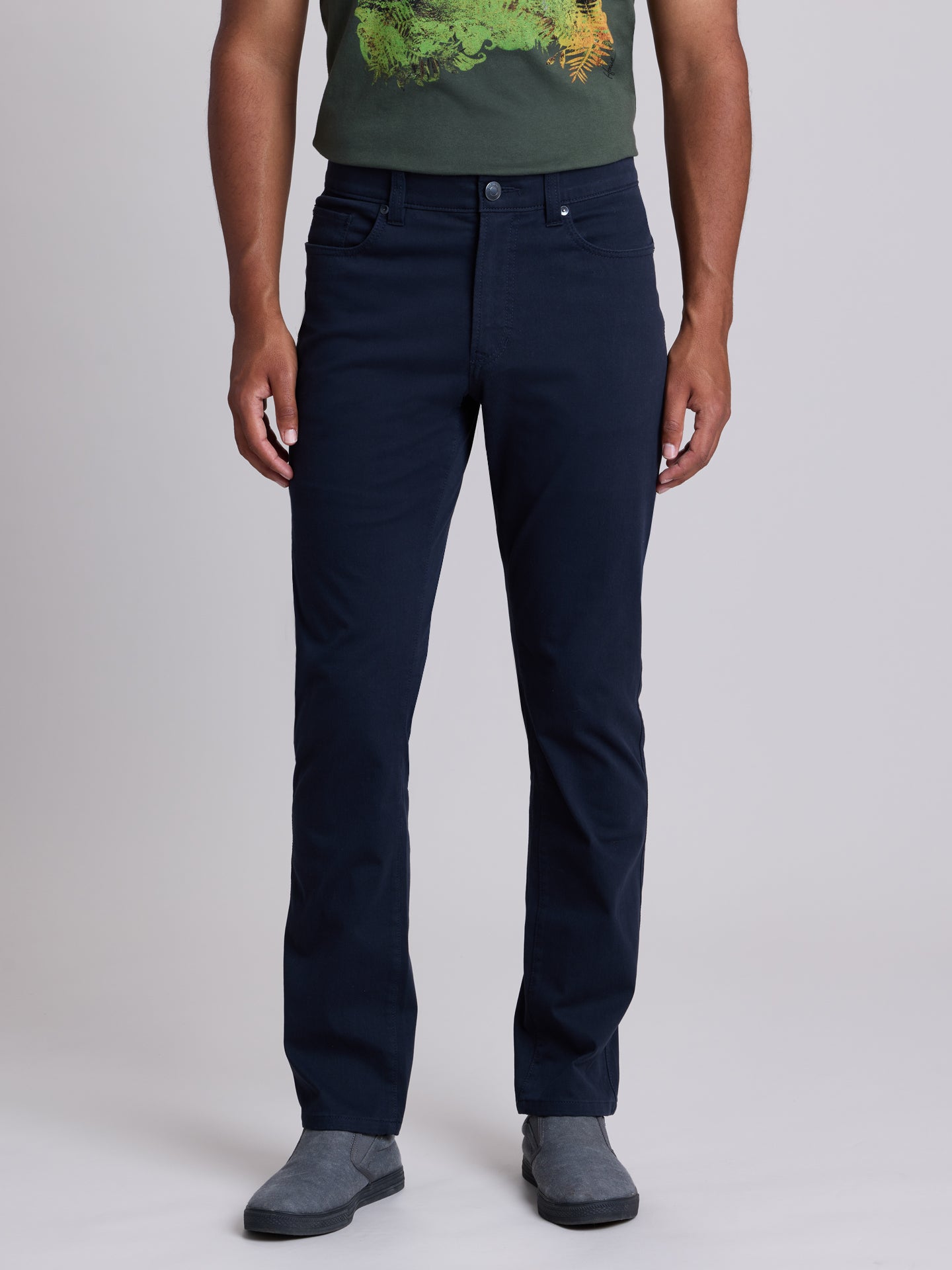 A man wearing dark navy blue straight-leg jeans, a green graphic t-shirt, and gray slip-on shoes, standing against a light gray studio background. | Twilight