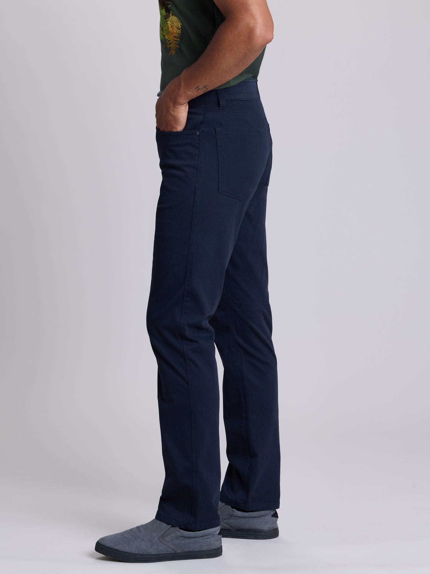 A side profile view of a person wearing dark navy blue trousers, a dark green t-shirt, and gray slip-on shoes, standing against a light gray studio background. | Twilight