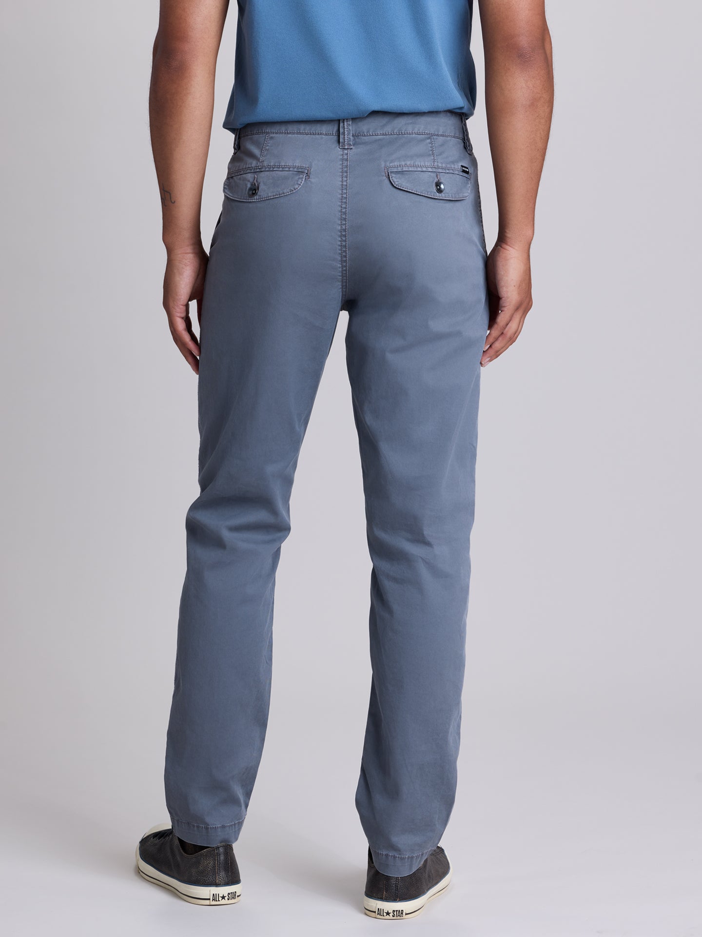 Rear view of a person wearing slate blue straight-leg chinos with a blue shirt and dark sneakers, photographed against a neutral gray studio  background. | Condor