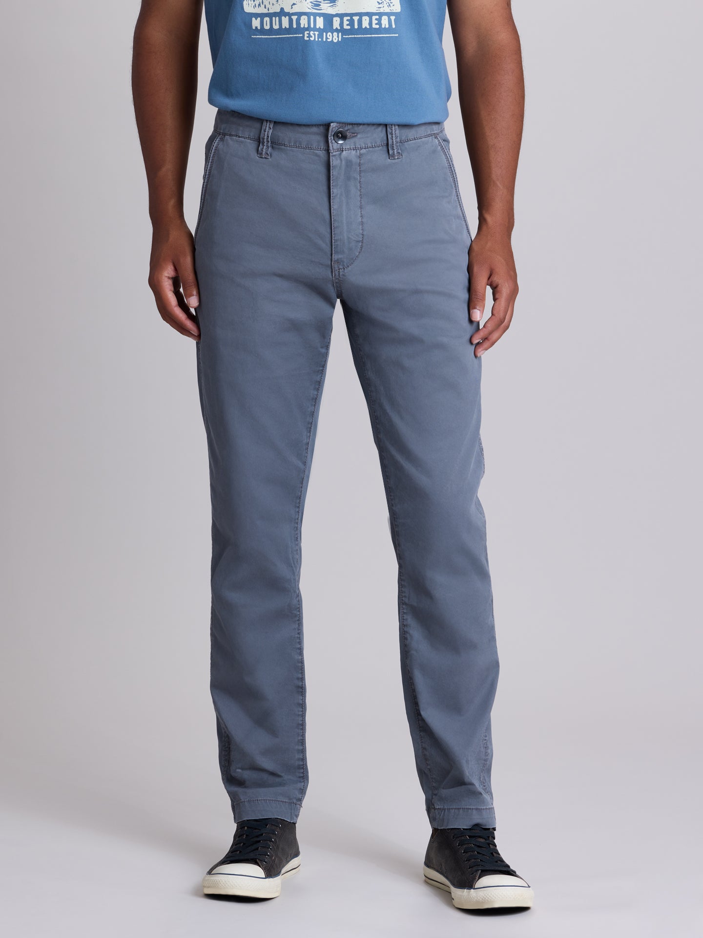 A person wearing slim-fit blue-gray chino pants, a blue graphic t-shirt, and black low-top sneakers, photographed against a light gray studio background. | Condor