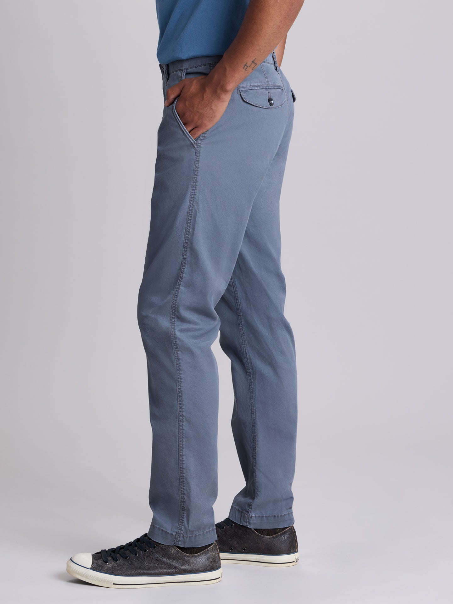A person shown from the waist down wearing a pair of slim-fit blue chinos with black sneakers, standing against a light gray studio background. | Condor