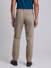 A man wearing beige straight-leg chino pants, a blue t-shirt, and brown leather shoes, shown from a rear three-quarter view against a light gray studio background. | Dugout