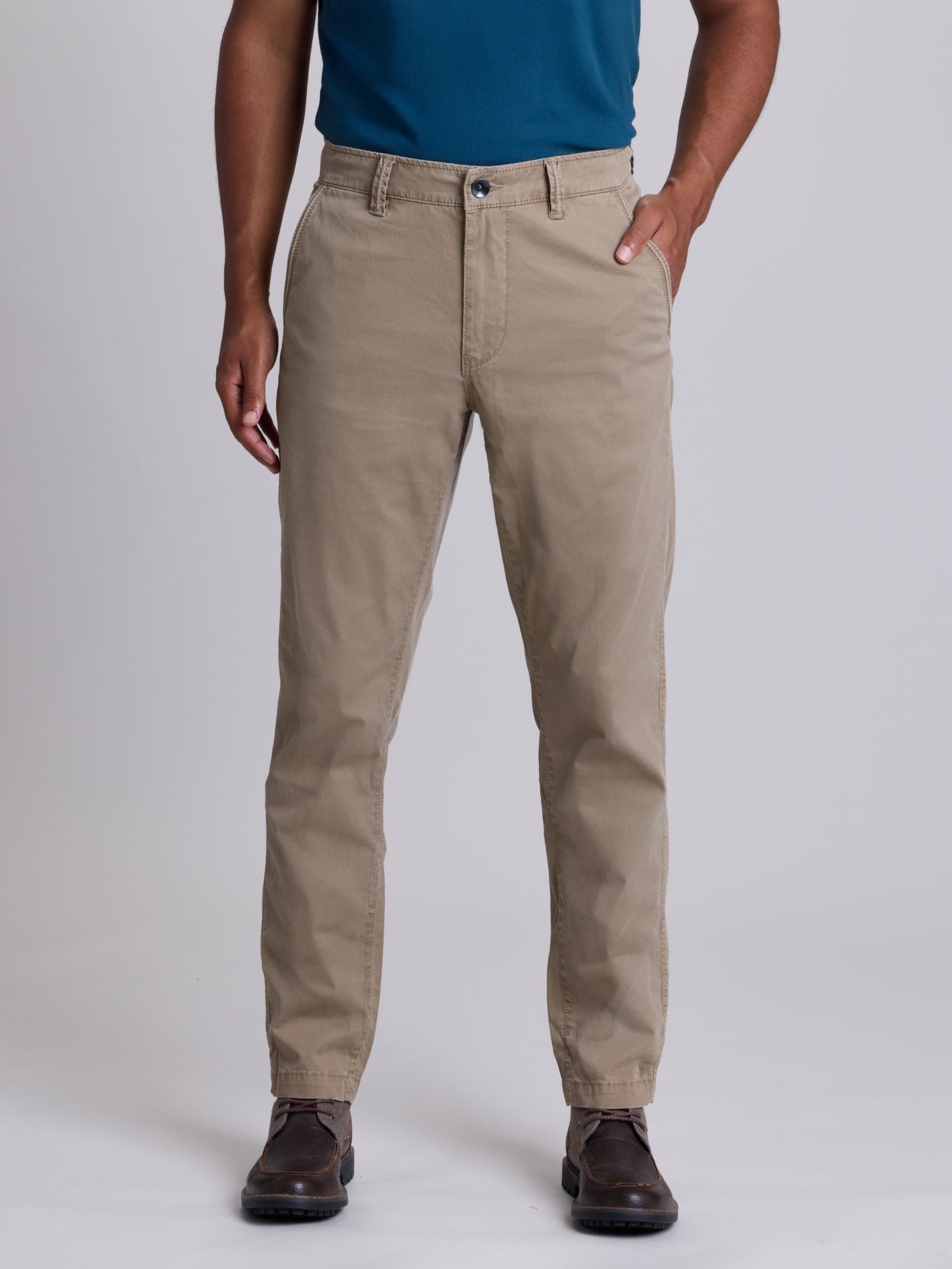 A man wearing khaki straight-leg chinos with a blue t-shirt and dark brown leather dress shoes, standing against a light gray studio background. | Dugout