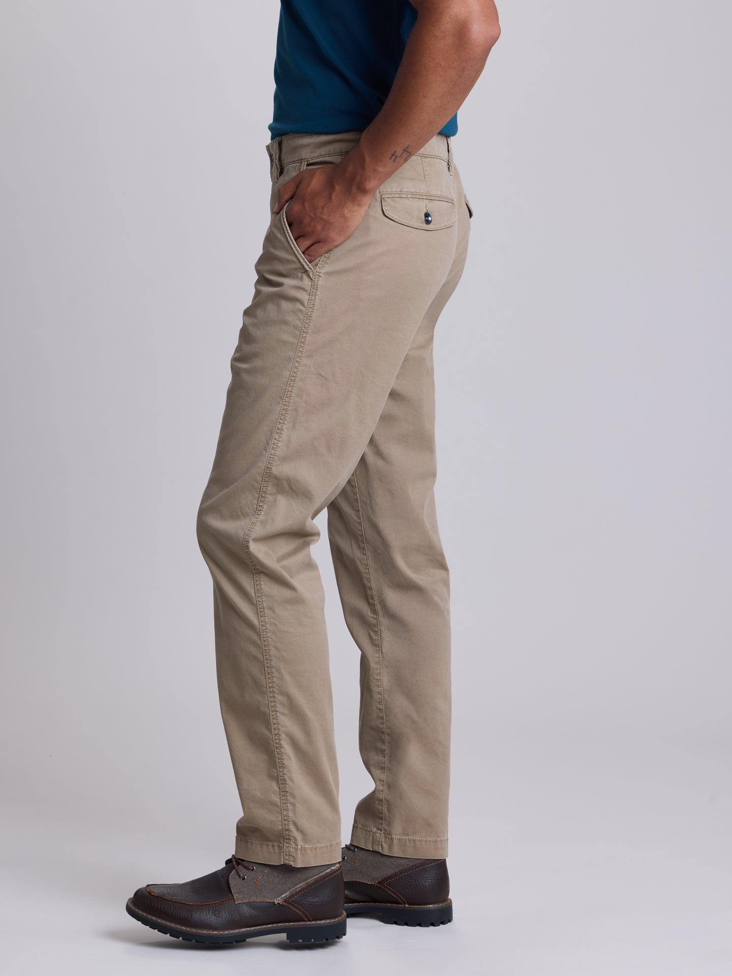 A side profile view of a person wearing beige chinos with a blue shirt and dark brown leather shoes, standing against a light gray studio background. | Dugout