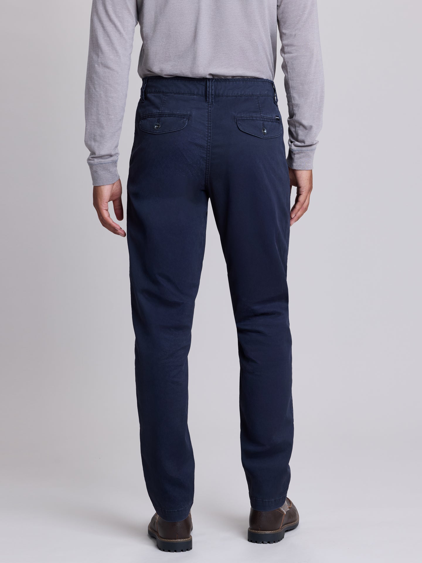 A man viewed from the back wearing navy blue trousers, a light gray long-sleeve shirt, and brown shoes, standing against a light gray studio background. | Twilight