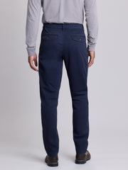 A man viewed from the back wearing navy blue trousers, a light gray long-sleeve shirt, and brown shoes, standing against a light gray studio background. | Twilight