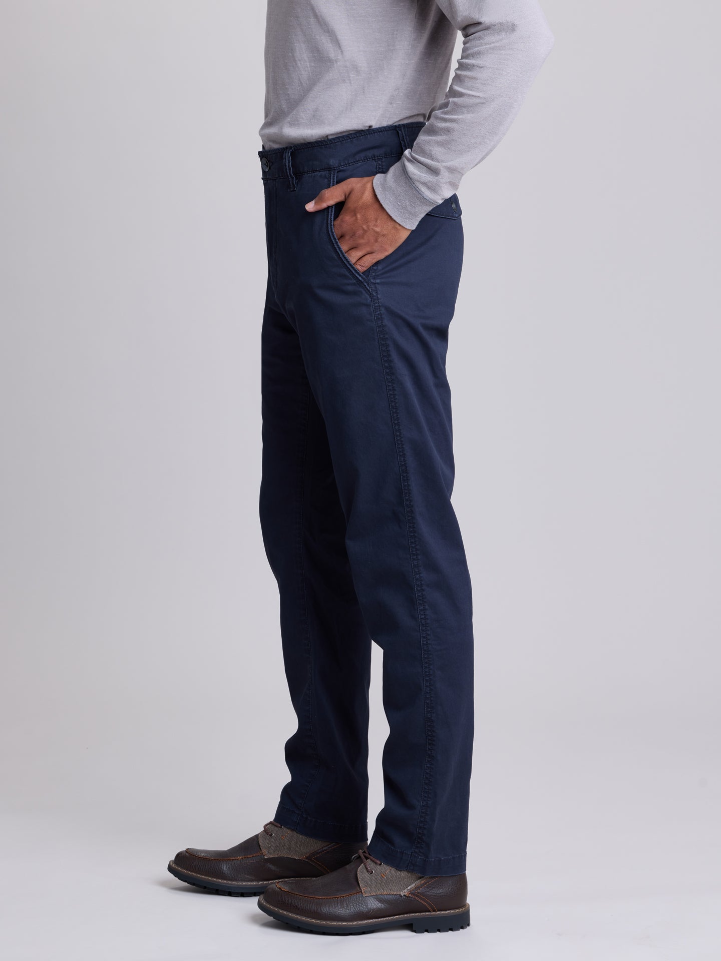 A person shown from the waist down wearing dark navy blue trousers, a light gray long-sleeve shirt, and brown leather shoes, standing against a light gray studio background. | Twilight