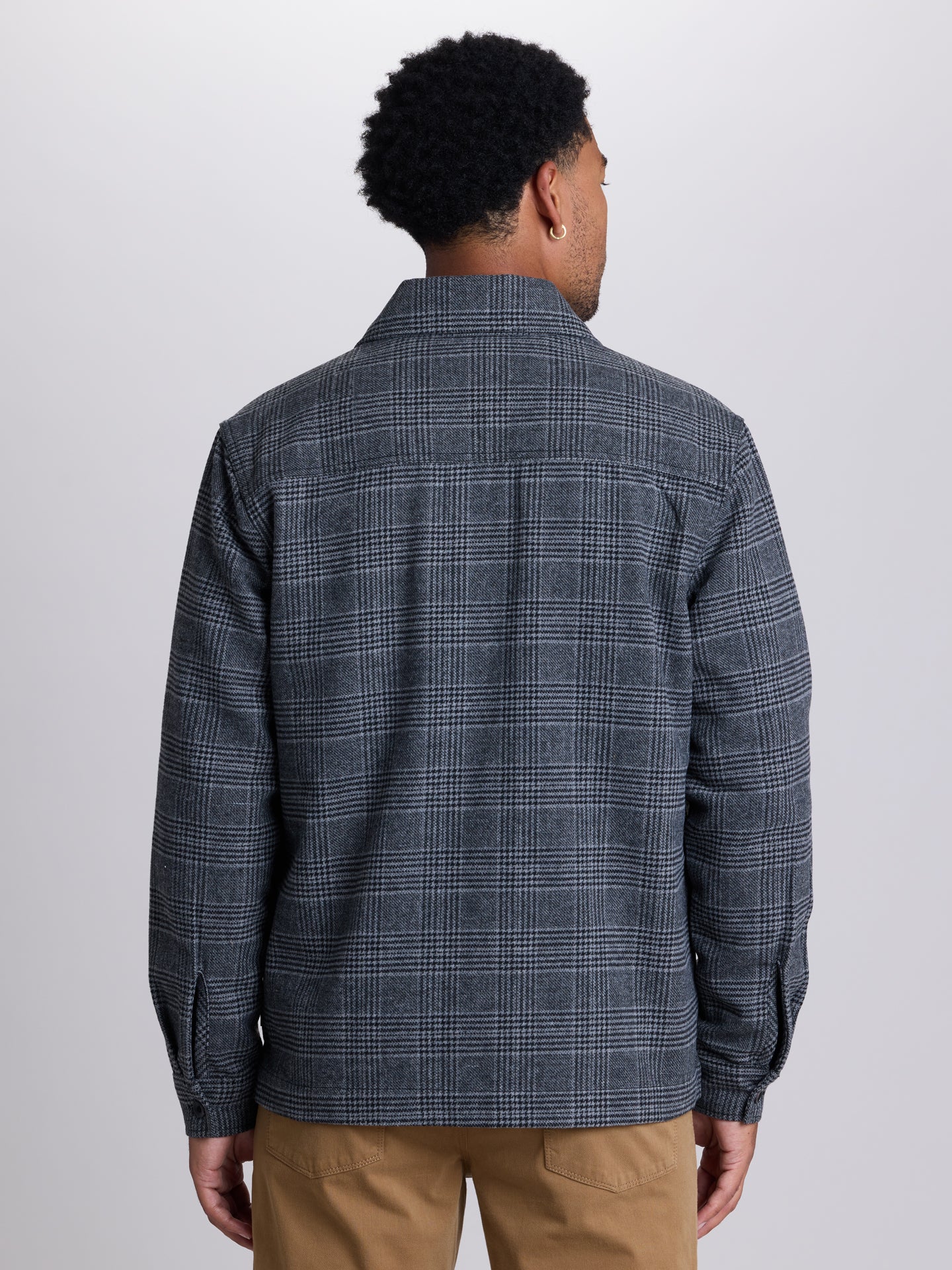 A man viewed from the back wearing a dark gray and navy plaid button-down shirt with tan pants, set against a plain light gray studio background. | Forged