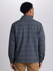 A man viewed from the back wearing a dark gray and navy plaid button-down shirt with tan pants, set against a plain light gray studio background. | Forged